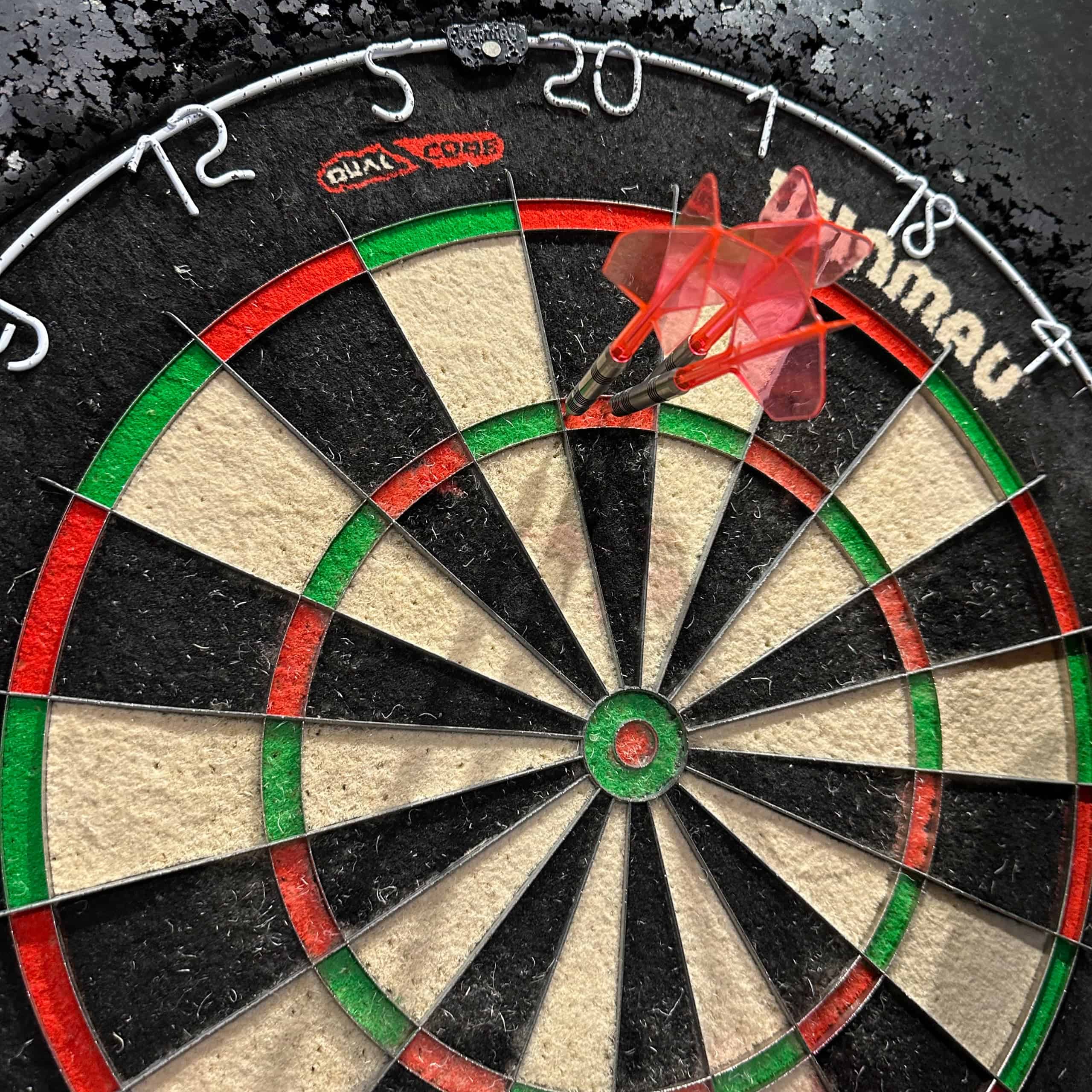 Three darts with translucent pinkish flights and shafts all stuck at triple 20 on a Winmau board.