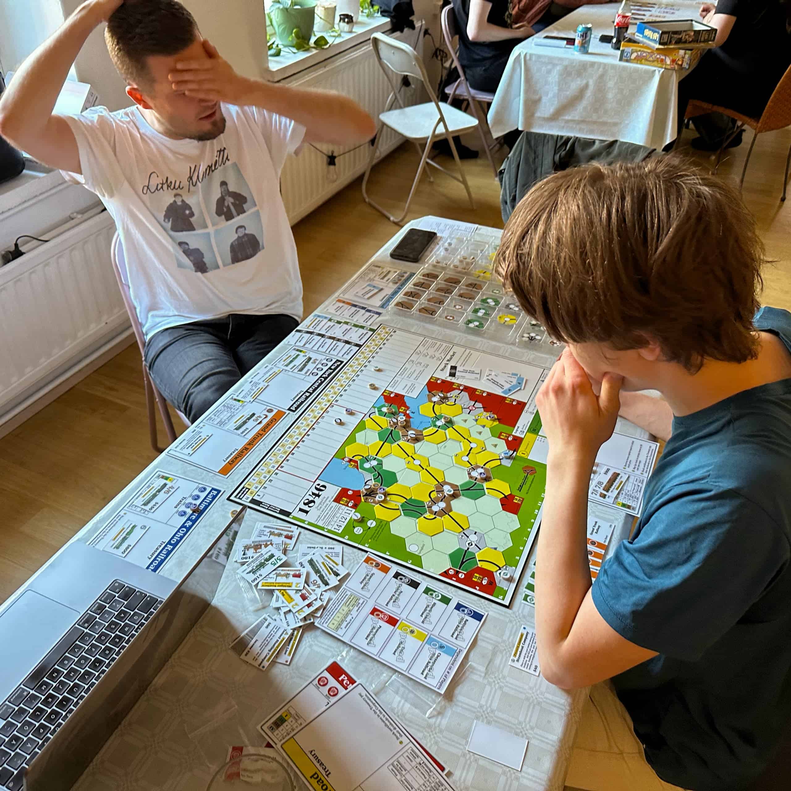 Two people sitting at the table where 1846 is played. One covers their eyes with their hand, the other leans on their hand, thinking. There's also a laptop on the table.