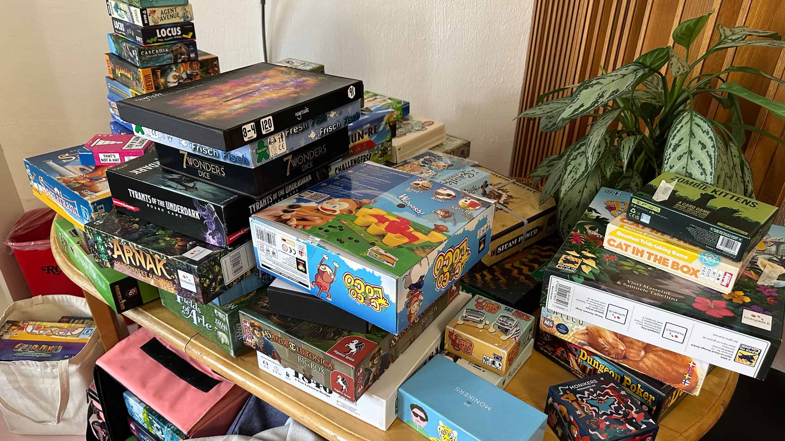 Piles of games on a table. Some games: Zombie Kittens, Cat in the Box, Monikers, String Railway, Coconuts, Rayroads, Fresh Fish, 7 Wonders Dice, Steam Power, Hansa Teutonica, Tyrants of the Underdark, Lost Ruins of Arnak, Agent Avenue, Locus, Cascadia Rolling Rivers, Samarkand, That's Not a Hat.
